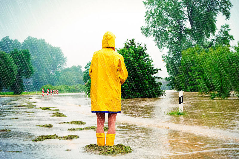 Eine Person steht mit gelben Regenmantel und Gummistiefeln bekleidet tief im Wasser auf einer Landstraße.